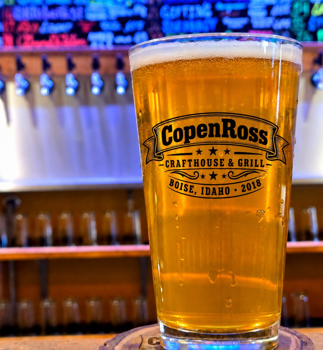 A pint of pale beer in a CopenRoss glass on a wood-slice coaster, with the tap wall and chalkboards behind